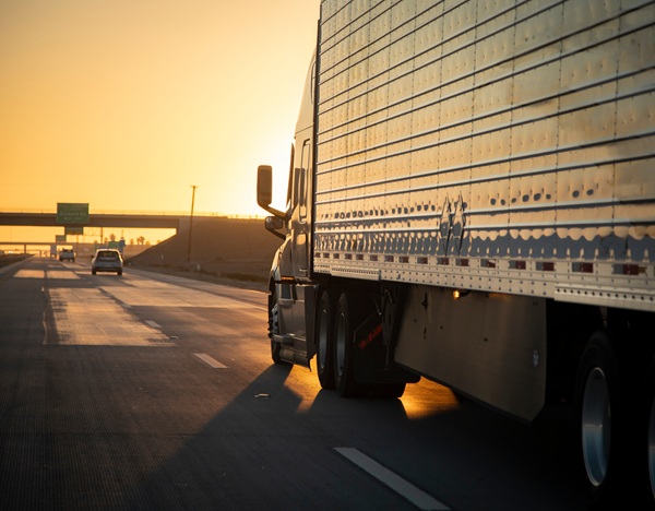 Semi-truck driving on the highway at sunset with sunlight reflecting off the trailer.