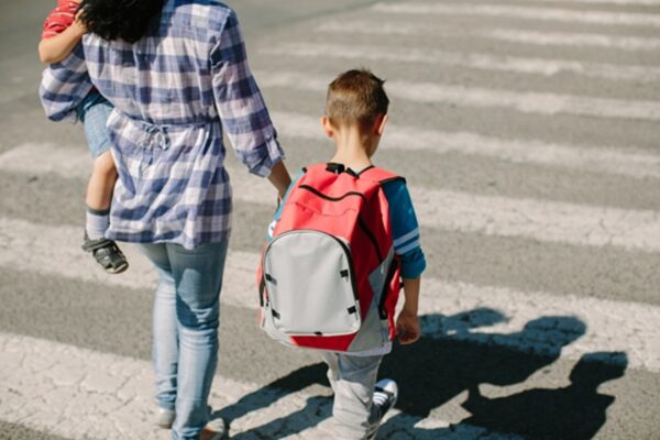 Parent walking across a crosswalk with a child wearing a red backpack and carrying another young child.