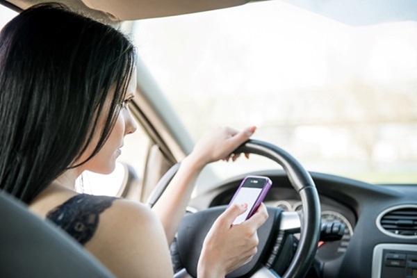 A young woman with long black hair holding a pink smartphone and looking down to text while driving a car.