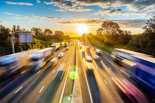 Heavy traffic of blurred semi-trucks and passenger cars moving along a multi-lane highway toward a bright sunset. (298379174)