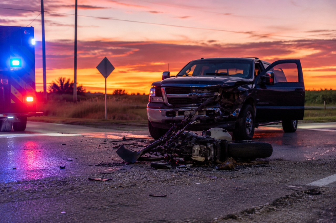 A severely damaged motorcycle lies on its side on a Texas road in front of a dark pickup truck with front-end damage and an open driver's door, with an emergency vehicle's flashing lights visible on the left under a dramatic sunset sky.
